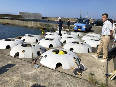 Installation of artificial reefs at a harbor. Several dome-shaped concrete reef units are arranged on the quay, with workers preparing and checking them, and the sea and work vehicles visible in the background.