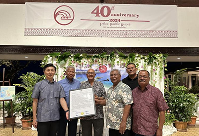 A commemorative photo taken at a ceremony venue. Several people stand in front of a banner reading 'Palau Pacific Resort 40th anniversary 2024', with a certificate of appreciation awarded by the Palau Senate and House of Delegates displayed at the center.