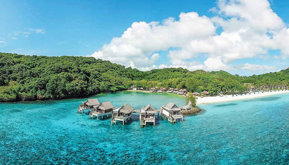 An overall view of Palau Pacific Resort surrounded by clear blue waters. Overwater bungalows extend into the sea, with a white sandy beach and lush green island forest in the background, showing a tropical resort integrated with the natural environment.