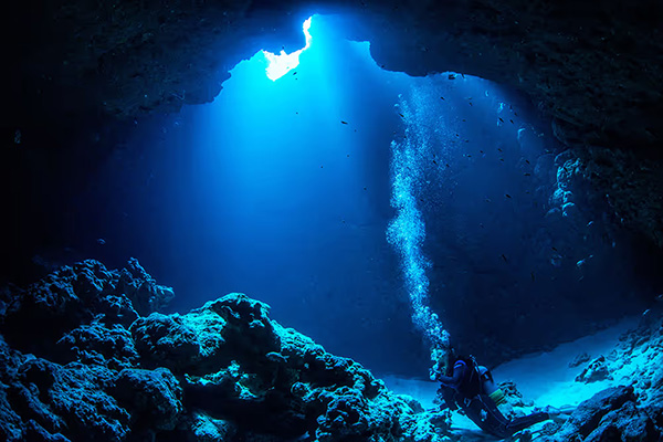 A diving spot inside an underwater cave. Blue light streams down from an opening above, illuminating the rocky seafloor as a diver swims through the cave. The scene conveys clear water and a mysterious underwater atmosphere.