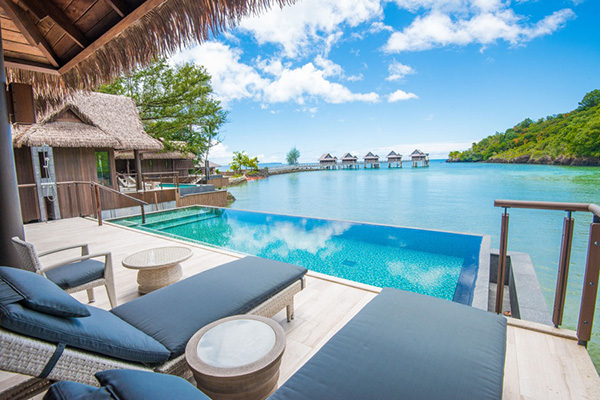 A view from the terrace of The Pristine Villas and Bungalows. An infinity pool and daybeds face the sea, with overwater bungalows visible across calm waters, conveying a luxury resort experience integrated with the natural surroundings.