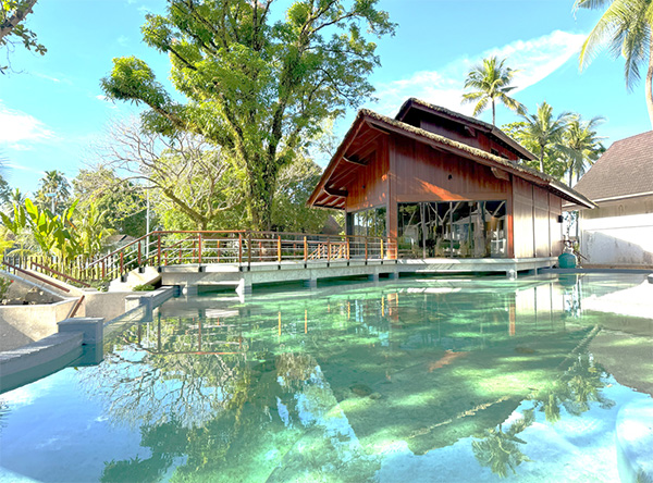 The exterior of Luke Nature Center. A wooden building stands beside clear water and is surrounded by lush greenery, with reflections of trees and the structure visible on the water surface, showing a nature-integrated experiential facility.