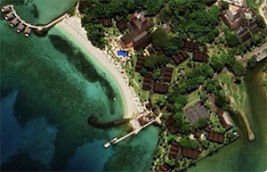 An aerial photograph from 2024 showing the area after development. A white sandy beach and piers line the coast, with resort buildings and pools set among lush greenery. The image illustrates the post-development landscape integrated with the natural environment.