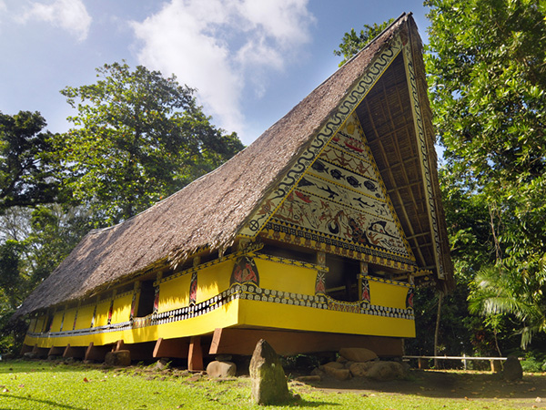 A traditional building known as an abai, used as a communal meeting house. It features a tall gabled roof, construction using wood and natural materials, and decorative patterns on the exterior walls. The structure is surrounded by lush forest, representing local culture and tradition.