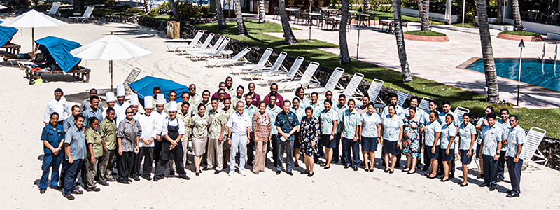 A group photo of hotel staff taken in an outdoor resort area. Many staff members stand together in rows with a sandy beach and palm trees in the foreground, and resort buildings and a pool visible in the background. The image conveys teamwork and a welcoming hospitality atmosphere.