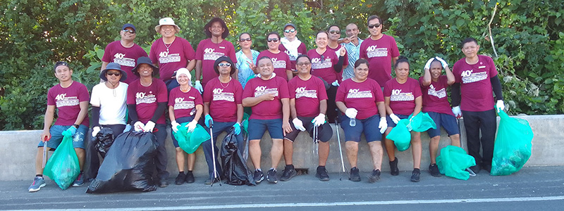 A group photo taken after a Green Care Committee activity. Many participants stand along a roadside, wearing gloves and holding bags of collected litter. Dense greenery in the background highlights an environmental cleanup and conservation effort.