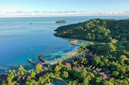 An aerial view of a resort site harmonized with nature. Piers and overwater facilities extend into clear blue waters, while low-rise buildings are nestled among lush green forests along the coast. The image shows a resort environment integrated with the surrounding natural landscape.