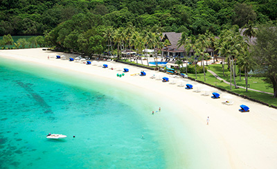 An aerial view of the beach after renovation. White sand stretches along the shore with neatly arranged beach umbrellas and lounge chairs, backed by palm trees, resort facilities, and lush forest. The scene shows a well-maintained coastal environment harmonized with clear turquoise waters.