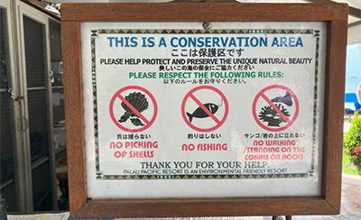 An informational sign for hotel guests about environmental protection. The sign states that the area is a conservation zone and shows rules such as no picking of shells, no fishing, and no walking or standing on corals or rocks, illustrated with icons and written in both Japanese and English. It encourages guests to help protect the marine environment.